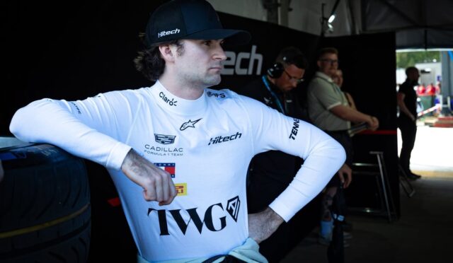 Race car driver in a white branded suit and black cap leans against a tire in a pit area, looking to the side.