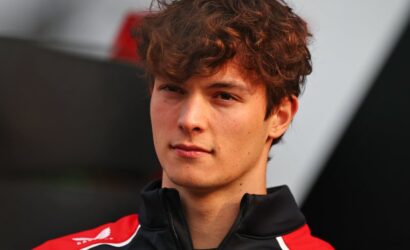 Young man with wavy brown hair wearing a red and black zip-up jacket, looking at the camera.