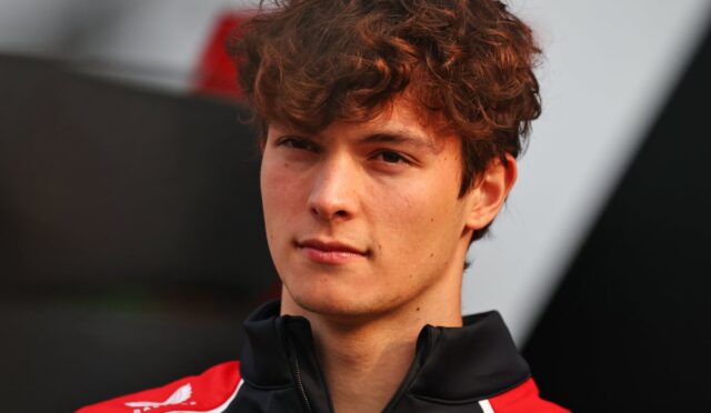 Young man with wavy brown hair wearing a red and black zip-up jacket, looking at the camera.