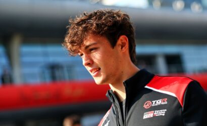 Young man with short, tousled brown hair wearing a black and red racing jacket at a race track, smiling slightly.
