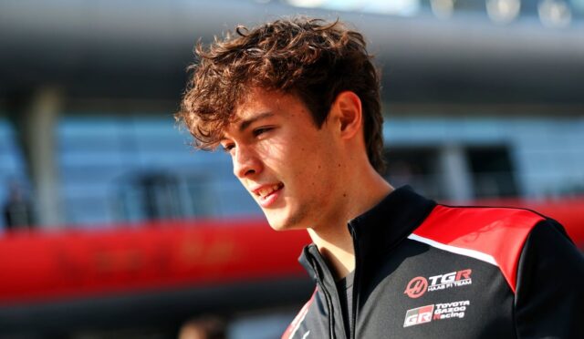 Young man with short, tousled brown hair wearing a black and red racing jacket at a race track, smiling slightly.