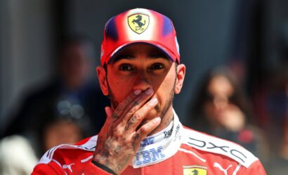 Portrait of a man in a red Ferrari cap, with his tattooed hand over his mouth during a moment at an event.