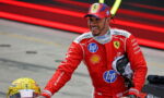 Ferrari Formula 1 driver in red racing suit smiling, reaching toward a yellow helmet on the pit lane, with sponsor logos visible on suit.