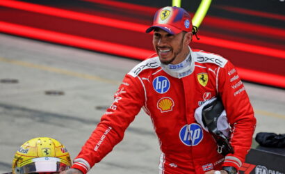 Ferrari Formula 1 driver in red racing suit smiling, reaching toward a yellow helmet on the pit lane, with sponsor logos visible on suit.