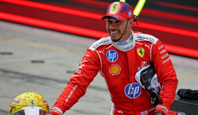 Ferrari Formula 1 driver in red racing suit smiling, reaching toward a yellow helmet on the pit lane, with sponsor logos visible on suit.