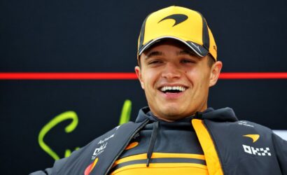 Young man in a yellow-orange cap and gray team jacket smiling at a race event.