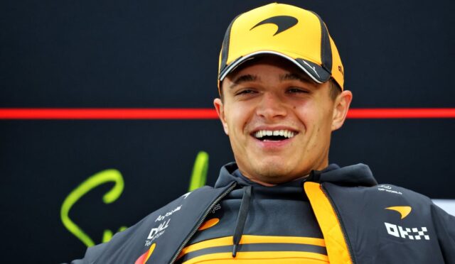 Young man in a yellow-orange cap and gray team jacket smiling at a race event.