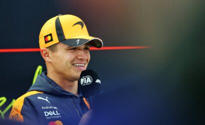 Male race driver in yellow cap and navy jacket, smiling during a televised interview with a microphone in front of him.