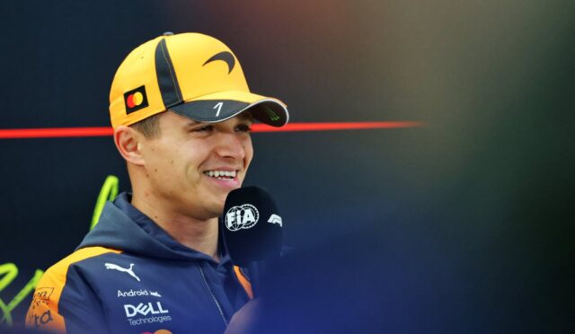 Male race driver in yellow cap and navy jacket, smiling during a televised interview with a microphone in front of him.