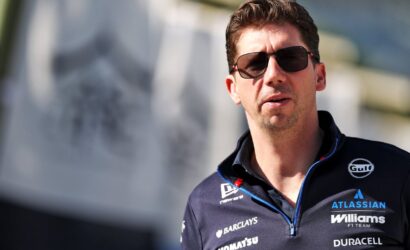 Man wearing sunglasses and a dark racing-team polo with sponsor logos, looking to the side.