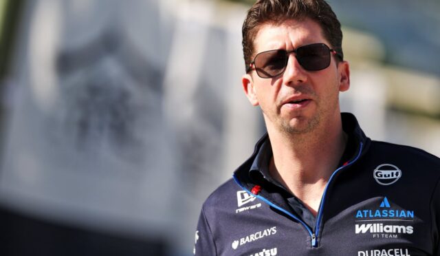 Man wearing sunglasses and a dark racing-team polo with sponsor logos, looking to the side.