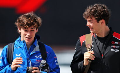 Two young men with backpacks smile and chat outdoors; one in a blue jacket holding a bottle and tumbler.