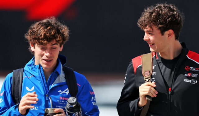 Two young men with backpacks smile and chat outdoors; one in a blue jacket holding a bottle and tumbler.