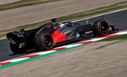 Formula race car in black and red livery speeds along a racetrack, with blurred background and red-white curbs visible in the foreground—action shot.