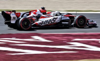 Race car in red, white, and black livery speeding on a race track with sponsor logos visible on the bodywork.
