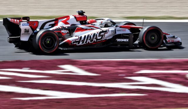Race car in red, white, and black livery speeding on a race track with sponsor logos visible on the bodywork.