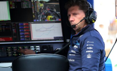 Race engineer wearing a headset sits at a console with multiple telemetry screens in a pit garage.