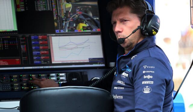 Race engineer wearing a headset sits at a console with multiple telemetry screens in a pit garage.
