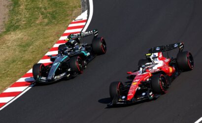 Two Formula 1 cars race side by side through a left-hand curve, a red Ferrari ahead of a dark Mercedes.
