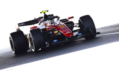 Red and black Formula 1 race car on a track, driver in white helmet navigating a corner, sponsor logos visible on the nose and sides