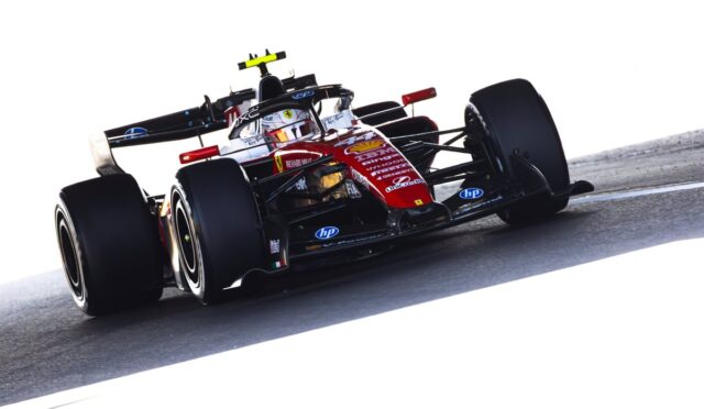Red and black Formula 1 race car on a track, driver in white helmet navigating a corner, sponsor logos visible on the nose and sides