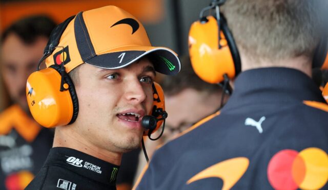Race car driver wearing an orange headset and cap, talking to a teammate in the pit area.