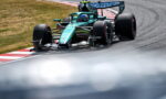 Teal Formula 1 race car cornering on a track, near red-and-white curbs with grass beside it.