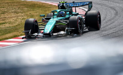 Teal Formula 1 race car cornering on a track, near red-and-white curbs with grass beside it.