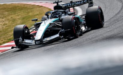 Mercedes Formula 1 car navigates a tight corner on a race track, tires gripping the red-and-white curbs.