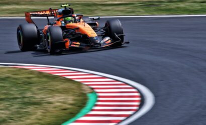 Orange Formula 1 car on a racetrack turning into a curve, speed blurred background shows motion.