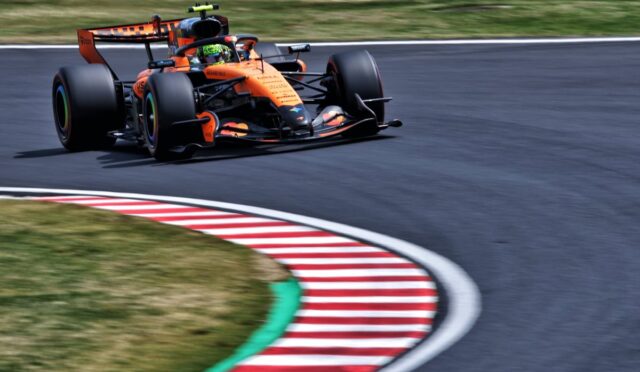 Orange Formula 1 car on a racetrack turning into a curve, speed blurred background shows motion.