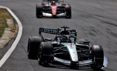 Mercedes F1 race car in silver/black livery navigating a curve on a race track, another red car following in the background