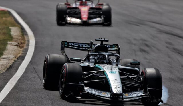 Mercedes F1 race car in silver/black livery navigating a curve on a race track, another red car following in the background