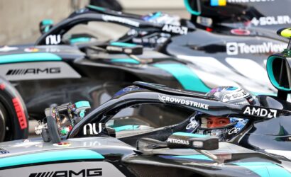 Close-up of a Formula 1 car cockpit with a driver in a helmet, surrounded by other branded race cars in the pit lane (AMD, CrowdStrike, Petronas).