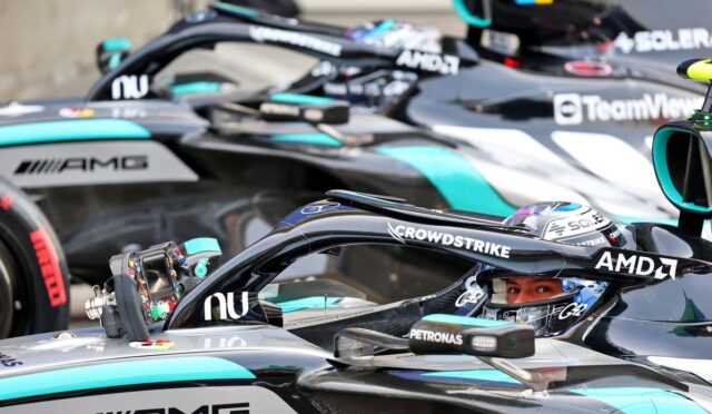 Close-up of a Formula 1 car cockpit with a driver in a helmet, surrounded by other branded race cars in the pit lane (AMD, CrowdStrike, Petronas).