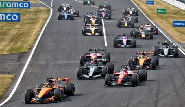 A pack of Formula 1 cars race on a track, close together at the start with sponsor boards along the sides.