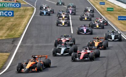 Group of Formula 1 cars at the start of a race on a track, with colorful liveries and sponsor boards along the sides.