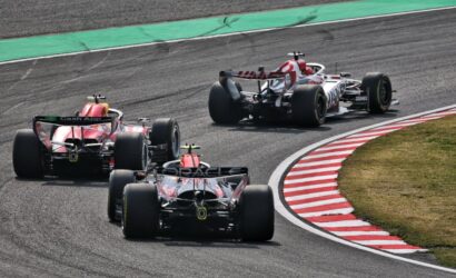 Three Formula 1 cars race through a tight corner on a track, tires smoking slightly and red-and-white curbs visible on the inside.
