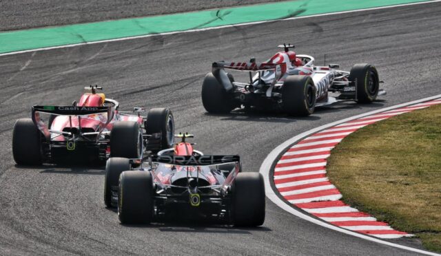 Three Formula 1 cars race through a tight corner on a track, tires smoking slightly and red-and-white curbs visible on the inside.