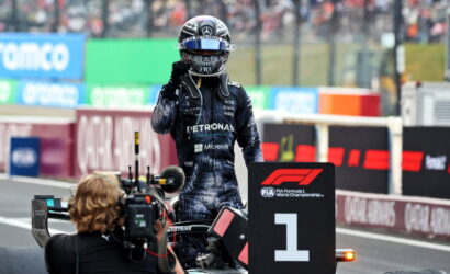 F1 winner in a Petronas Mercedes suit stands beside the No. 1 podium marker as a cameraman shoots the moment on the track side barriers in the background.