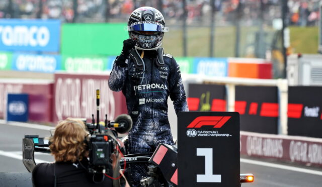 F1 winner in a Petronas Mercedes suit stands beside the No. 1 podium marker as a cameraman shoots the moment on the track side barriers in the background.