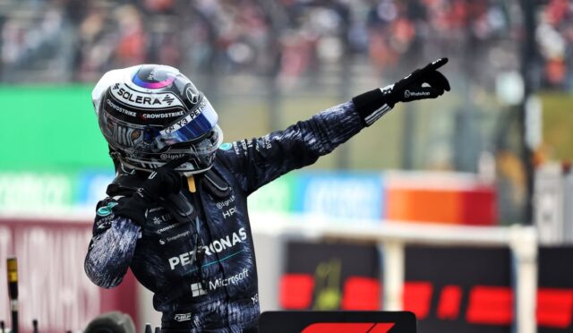 F1 driver in Petronas suit with helmet pointing toward the crowd on a race track stride, surrounded by pit area.