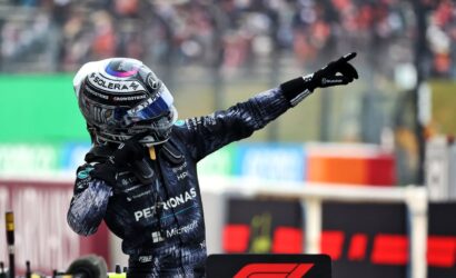 Racing driver in a dark Petronas racing suit and helmet raises a gloved arm and points toward the track in a pit-area with a blurred crowd behind.