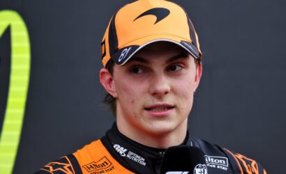 Young race car driver in orange cap and racing suit speaking into a microphone during a post-race interview.