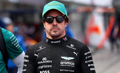 Man in a black Formula One racing suit with sponsor logos, teal cap and sunglasses, in a pit area.