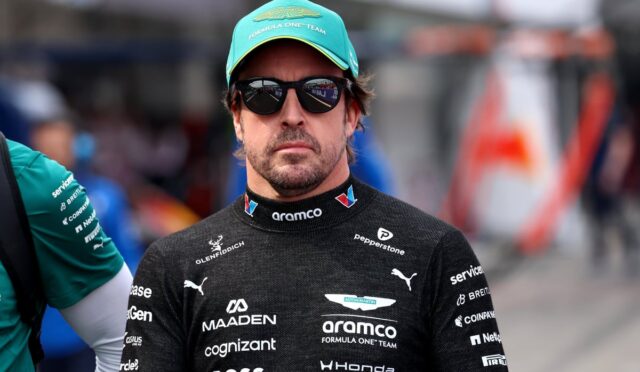 Man in a black Formula One racing suit with sponsor logos, teal cap and sunglasses, in a pit area.