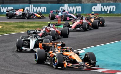 F1 cars in a tight corner on a race track, orange McLaren leading, with Allwyn banners in the background.