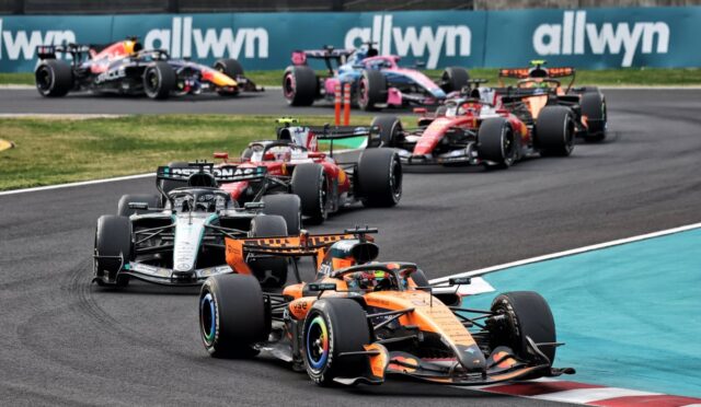 F1 cars in a tight corner on a race track, orange McLaren leading, with Allwyn banners in the background.