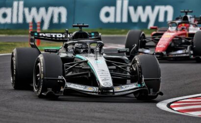 Mercedes Formula 1 car leading a race on a track with another red car close behind, blue allwyn barrier in the background.
