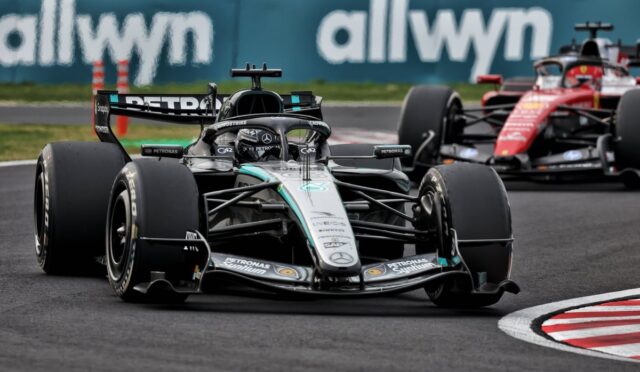 Mercedes Formula 1 car leading a race on a track with another red car close behind, blue allwyn barrier in the background.